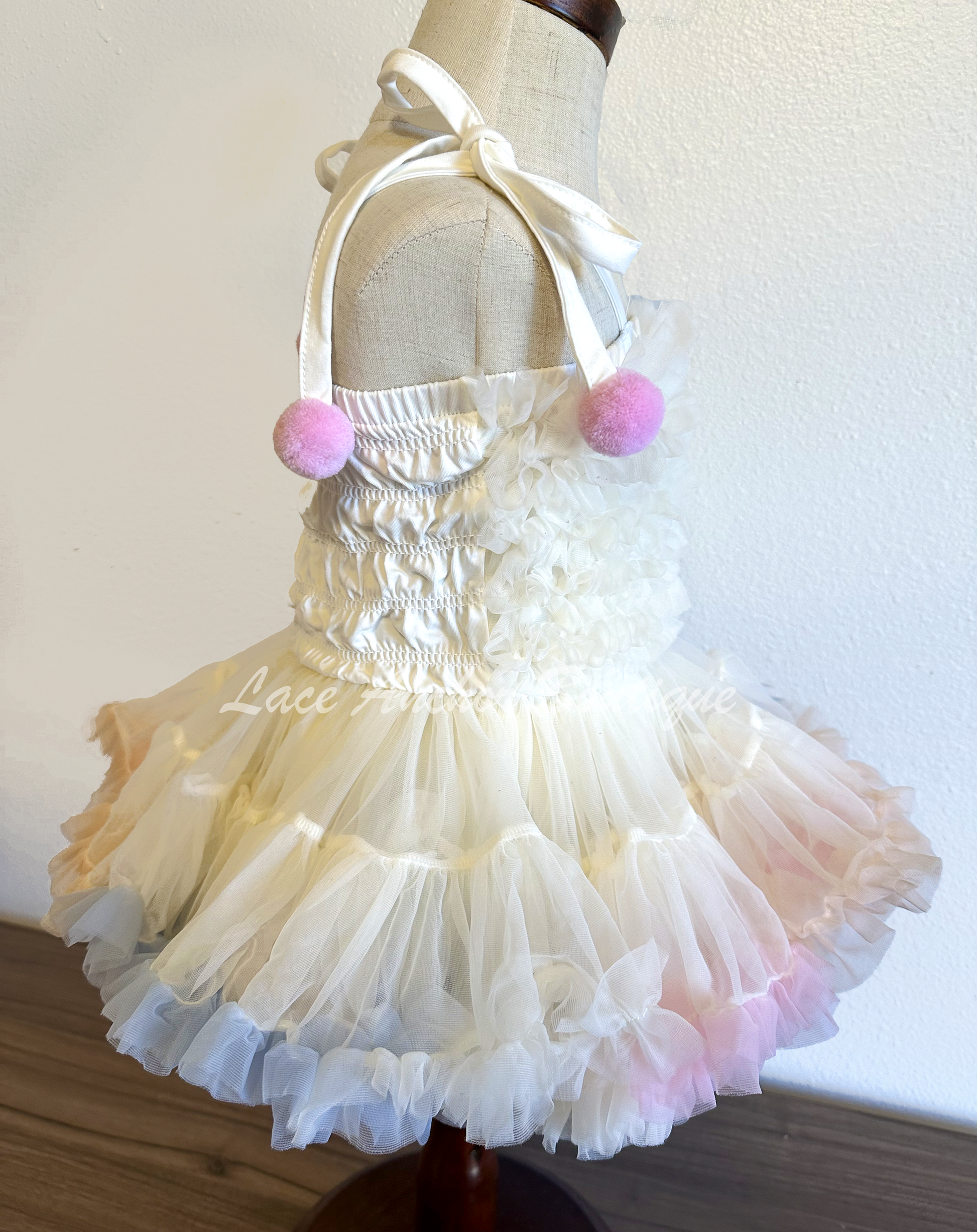 toddler baby girls pastel rainbow multi colored dress with pearl details, smocked ruffle top, and ruffle trim and tied pom pom straps. Unicorn colored theme birthday girl princess dress.