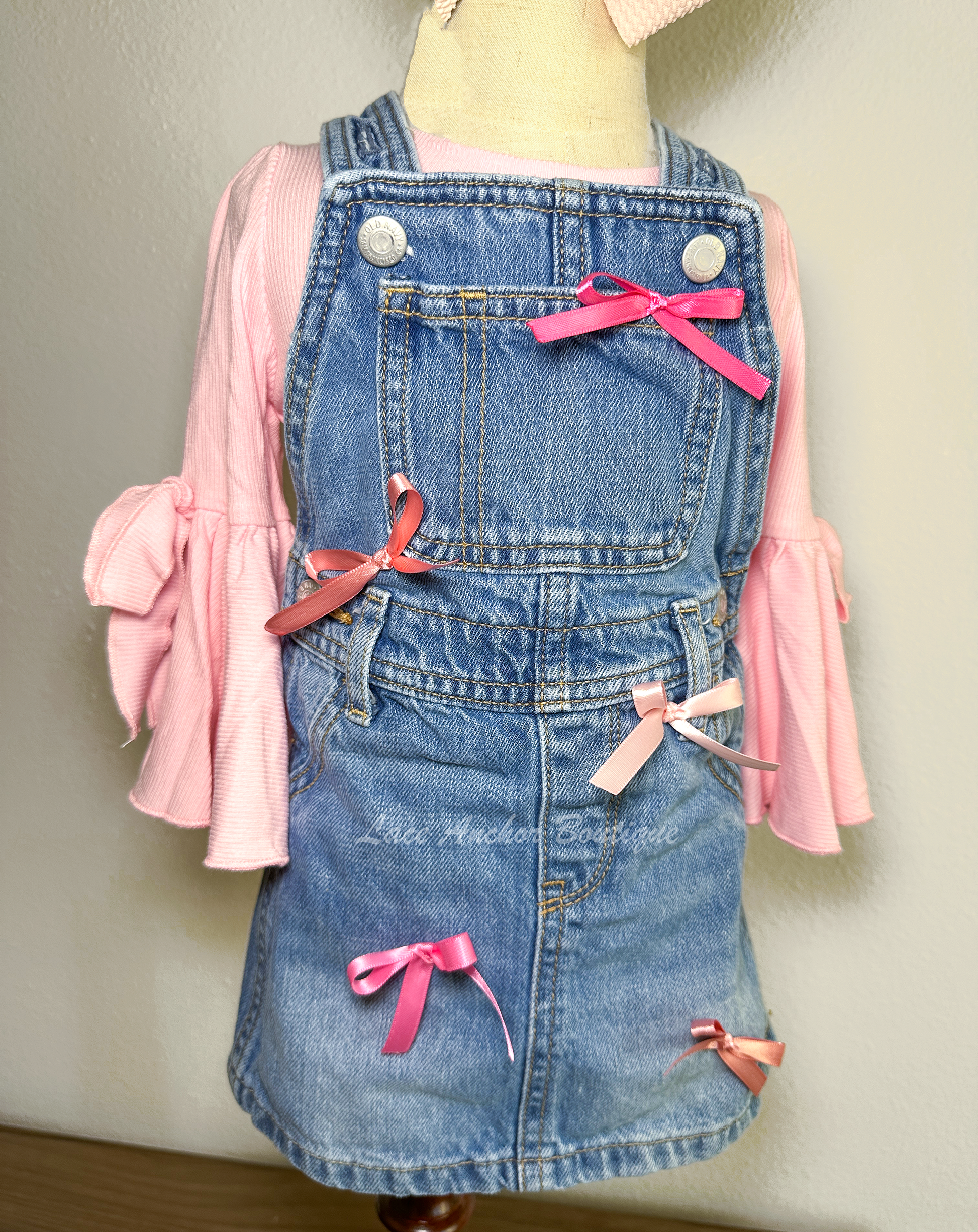 toddler girls blue denim overalls in a skirt style with multi colors of pink silky bows on a mannequin