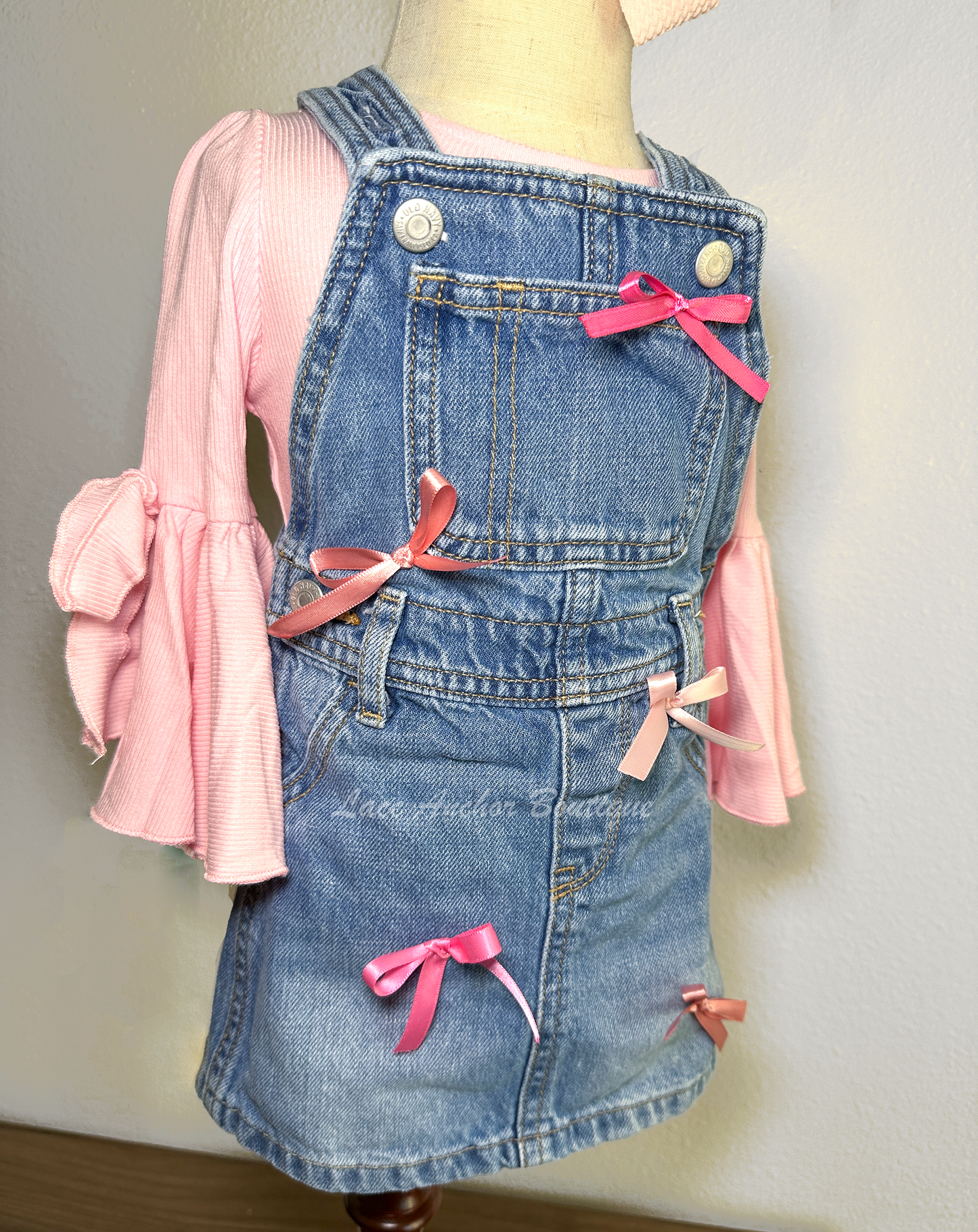 toddler girls blue denim overalls in a skirt style with multi colors of pink silky bows on a mannequin