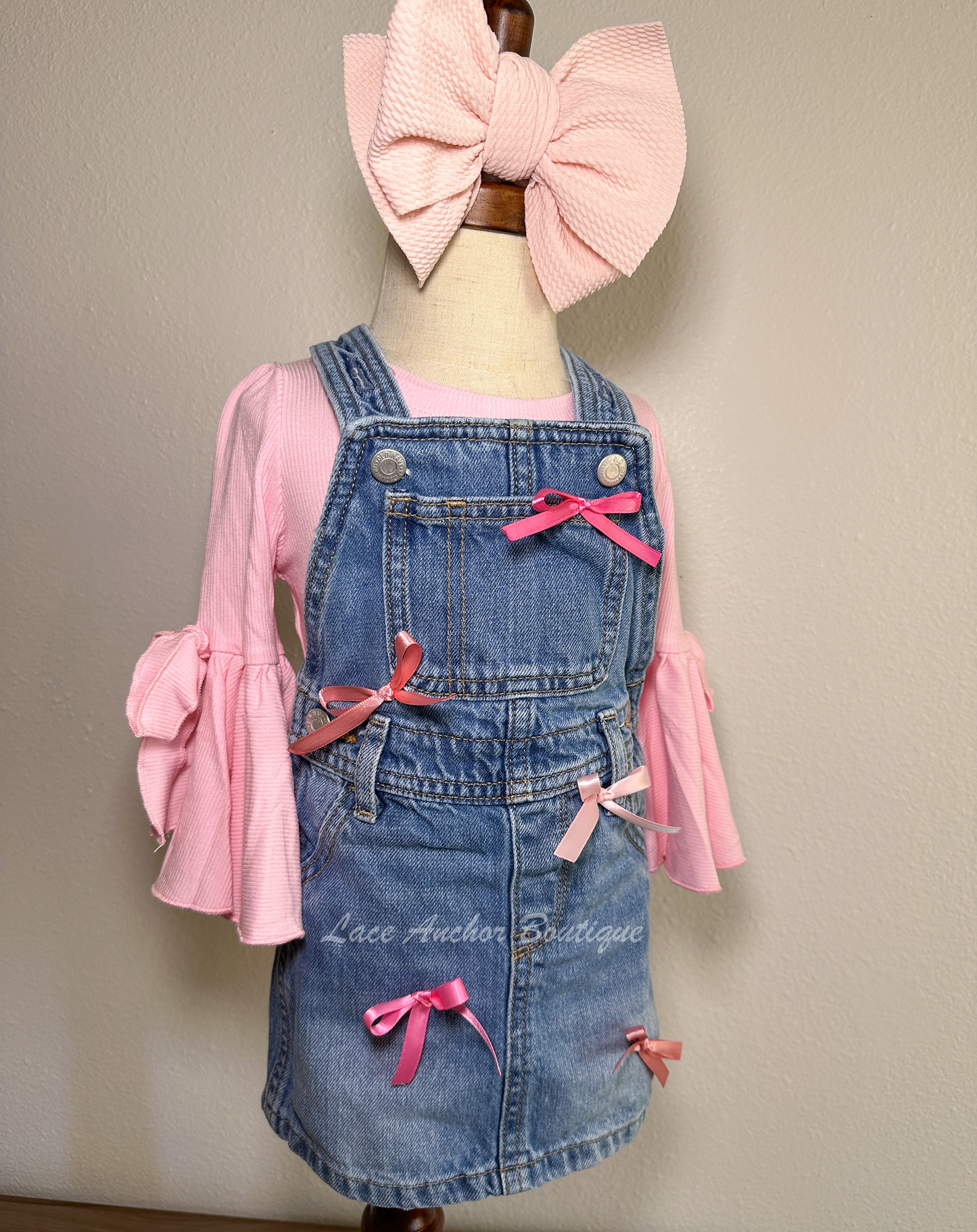 toddler girls blue denim overalls in a skirt style with multi colors of pink silky bows on a mannequin