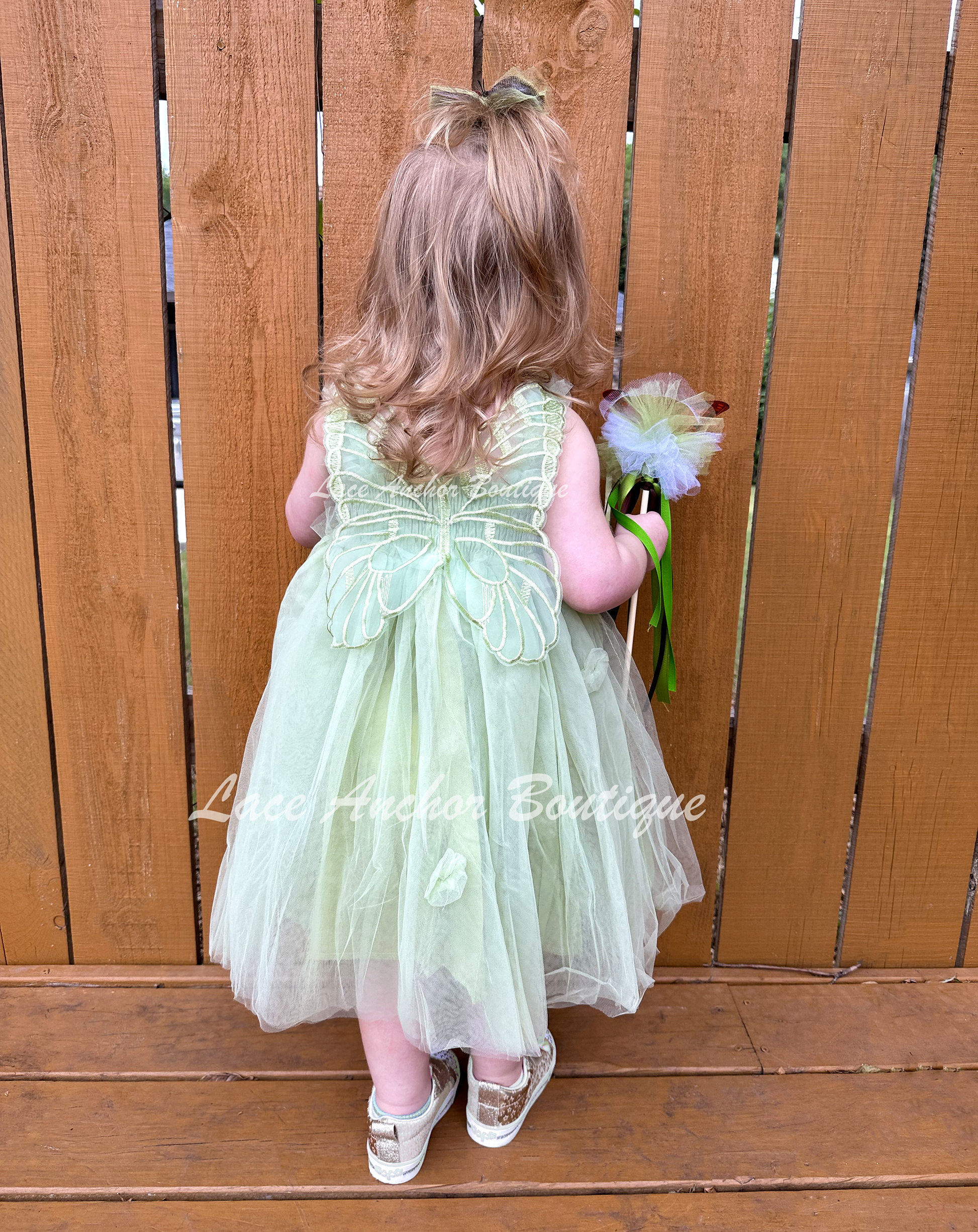 Sage Green tulle fairy girls dress with smocked top and tulle flowers on skirt. Attached butterfly wings and ruffled straps. Worn by toddler model.